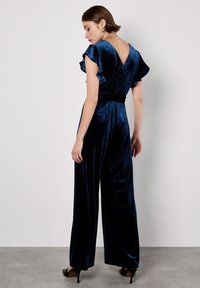 Mujer vestida con un mono de terciopelo azul marino con mangas acampanadas y espalda en V, de pie contra una pared blanca lisa con tacones negros.