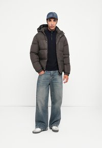 Mørk pufferjakke med hætte, navy zip-neck sweater, vide blege denim jeans og grå sneakers med hvide detaljer. Afslappet outfit.