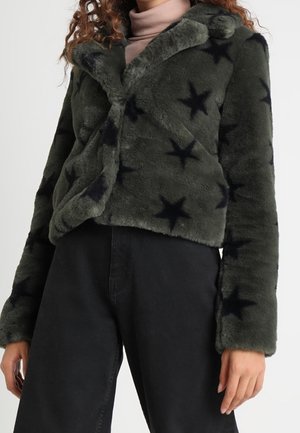 Veste en fausse fourrure avec des motifs étoiles noirs, portée sur un col roulé rose clair, assortie à un pantalon noir taille haute.