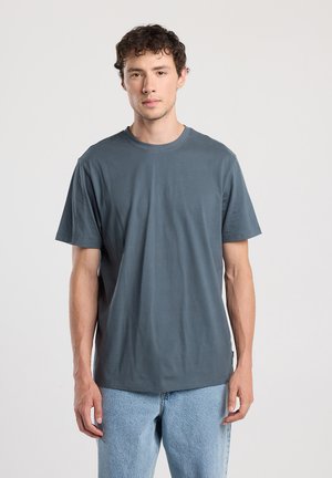 Young man with short curly hair wearing a plain dark gray t-shirt and light blue jeans, standing against a neutral background.