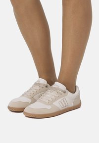 Person trägt beige-weiße Schnürsneaker mit Gummisohlen und steht vor einem schlichten weißen Hintergrund.