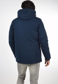 Temno modra pufasta jakna s kapuco, ki ima gladko teksturo, elastične manšete in ravno obrobo. Nosite jo z svetlo sivo jeans hlačami.