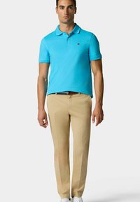 Tyrkysové polo tričko s límečkem, krátkými rukávy a malým logem na hrudi, kombinované s béžovými kalhotami a bílými teniskami.