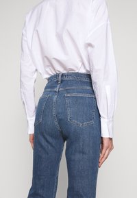 Jeans de denim azul claro con cintura alta, que cuentan con cinco bolsillos y un patrón de costura clásico. Llevados bajo una camisa blanca de corte holgado.