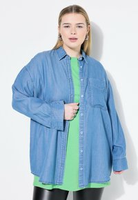 Camicia di denim azzurro chiaro con taschino frontale, design con bottoni, maniche arrotolate, sovrapposta a una maglietta verde, indossata con leggings neri lucidi.
