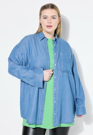 Lichtblauwe denimshirt met een borstzak, knoopsluiting, opgerolde mouwen, over een groen T-shirt gedragen, gecombineerd met glanzende zwarte leggings.