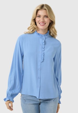Himmelblaue Bluse mit Rüschensaum am Kragen, Knopföffnung und langen Ärmeln, die an den Bündchen gerafft sind, aus glattem Stoff. Kombiniert mit Jeans.