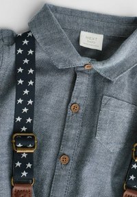Denimhemd mit Kragen, braunen Holzknöpfen und einer Brusttasche. An den Trägern befindet sich ein Sternmuster auf dunklem Hintergrund.