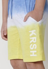 Bavlnené šortky v zmesi s modro-žltým ombre dizajnom, obsahujúce bočnú kapsu a výrazné biele písmo "KRSH" na žltej časti.