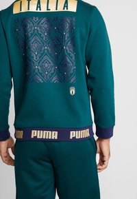Tyrkysová mikina s zlatým logem "ITALIA", složitě vzorovaná tmavě modrá záda a "PUMA" pás na spodní části v purpurové a zlaté barvě.
