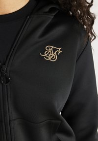 Zwarte ritsjas met een gestructureerde afwerking en een beige geborduurd logo op het borstgebied; heeft een hoge kraag en zijzakken.