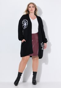 Černý pletený kardigan s bílou květinovou výšivkou, dlouhé rukávy, otevřený přední díl, boční kapsy, nošený přes bílý top a burgundskou sukni. Černé kotníkové boty.