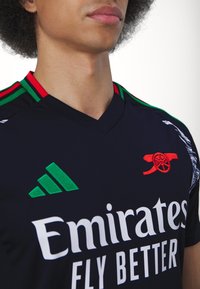 Jeune personne portant un maillot de sport sombre avec des rayures rouges et vertes, logo Adidas, emblème du canon d'Arsenal et texte "Emirates Fly Better".