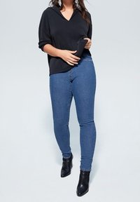 Nainen, joka pitää mustaa hupparia, sinisiä leggings-housuja ja mustia nilkkureita, pitää yllään hupparia vyötärön kohdalta yksiväristä taustaa vasten.