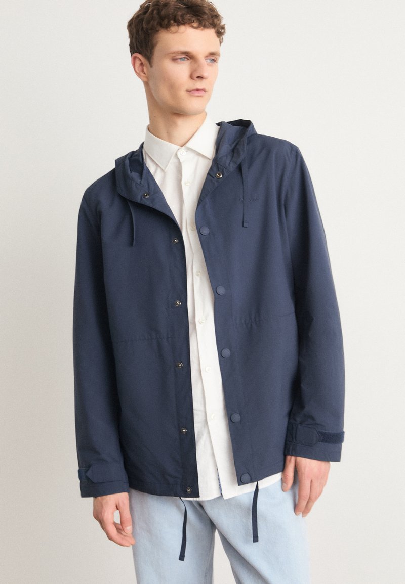 Navy blue hooded jacket made of lightweight fabric, featuring snap buttons, drawstring details, and a relaxed fit, worn over a white shirt.