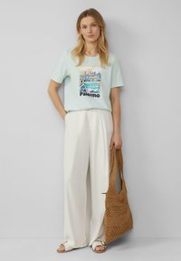 Licht mintgroen T-shirt met een Palermo-grafiek, gecombineerd met witte wijde broek. Gecombineerd met een bruine gevlochten tas en platte sandalen.