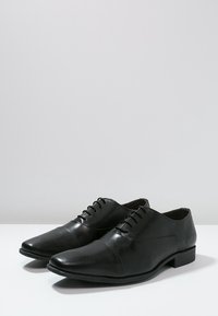 Chaussures habillées en cuir noir avec un nez pointu, texture lisse, design à lacets et un petit talon carré. Style minimaliste sans accents visibles.