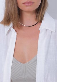 Collier ras du cou en perles noires et blanches porté sur un haut gris clair, associé à une chemise blanche boutonnée, avec un focus sur le col.