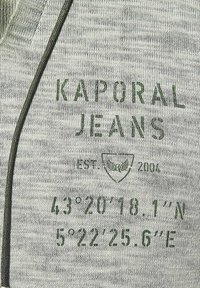 Grijs stof met een gemêleerde textuur. Bevat groene bedrukte tekst: "KAPORAL JEANS," "EST. 2004," en geografische coördinaten.