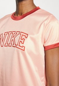 Närbild av en person som bär en blanka rosa t-shirt med röda ribbade kragar och ärmslut, med fet röd "NIKE"-text över bröstet.