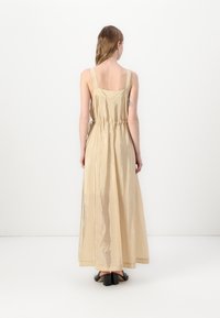 Duga, bez rukava bež haljina sa sjajnom teksturom, kvadratnim leđima i nabranim strukom. Nosena uz crne cipele s blok petom.