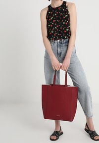 Červená kožená taška přes rameno s dvěma uchy, obdélníkový design, hladká textura a decentní logo. Modelka má na sobě květinový top a světlé džíny.