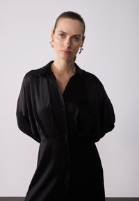 Rochie din satin negru cu nasturi în față, guler structurat și mâneci supradimensionate. Textură netedă și design minimalist.