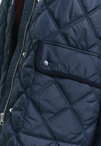 Marineblaue gesteppte Jacke mit Reißverschluss und Taschen mit Druckknopf. Verfügt über eine Diamantmuster-Steppung und kontrastierende Einfassung an der Tasche.