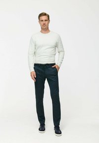Helles graues Langarmshirt, kombiniert mit dunkelblauen Hose. Das Model trägt marineblaue Sneakers. Einfaches, tailliertes Design mit glatter Textur.