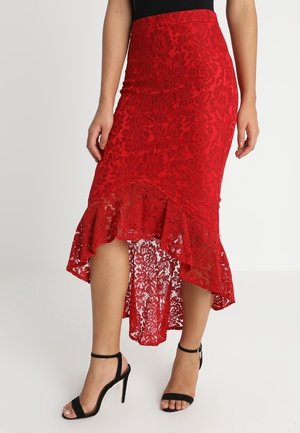 Femme portant une jupe en dentelle rouge à coupe asymétrique avec ourlet à volants et sandales à talons hauts noires avec bride à la cheville sur fond blanc.