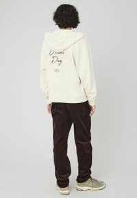 Sweat à capuche crème avec une grande capuche, poche avant et le texte "Creative Day" au dos. Associé à un pantalon en velours côtelé marron et des chaussures de sport.