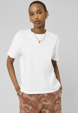 Femme portant un t-shirt blanc uni à manches courtes, un pantalon marron avec une broderie florale blanche, et des bijoux en or, debout les mains dans les poches.