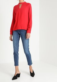 Blusa rossa a maniche lunghe con scollatura a chiave, abbinata a jeans aderenti blu con strisce laterali e scarpe con tacco nere.