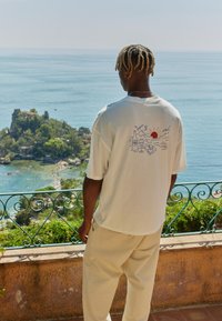 Hvit oversized T-skjorte med en fargerik grafikk av en sol, drikker og en bok på baksiden; brukt med beige bukser foran en kystutsikt.