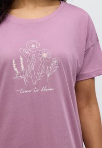 Λιλα κοντομάνικη μπλούζα με λευκό floral σχέδιο και τη φράση "time to bloom" με έναν χαλαρό, στρογγυλεμένο γραμματοσειρά.
