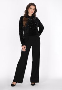 Sort fluffy sweater med farverige juveldetaljer ved halsudskæringen, parret med sorte vide bukser og sorte højhælede sko.
