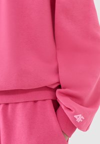 Gros plan d’une personne portant un sweat-shirt rose et un pantalon avec des poignets côtelés, montrant un petit logo "4F" sur le poignet de la manche.