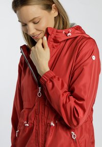 Rote, leichtgewichtige Jacke mit Kapuze, elastischen Bündchen und Kordelzug in der Taille. Mit einem Reißverschluss vorne und zwei Fronttaschen. Glatte Textur.