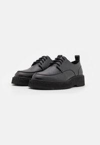 Scarpe in pelle nera con una finitura liscia, punta arrotondata e suola testurizzata sollevata. Presentano sei asole e un piccolo anello posteriore.