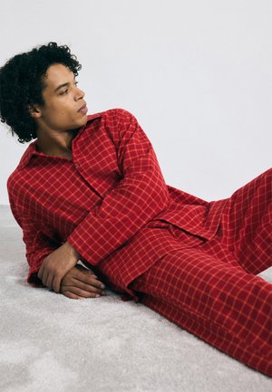 Rood geruit pyjamapak gemaakt van zachte stof, met een overhemd met knopen en lange mouwen en bijpassende broek, gelegd op een lichtgrijze ondergrond.