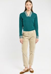 Haut à manches longues teal avec un col et des poignets à motifs, associé à un pantalon beige ajusté et des chaussures plates marron. Tenue simple et décontractée.