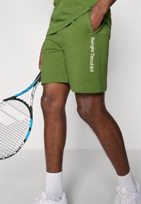 Calções de algodão verdes com bolsos laterais, apresentando uma impressão do logótipo em branco. Combinados com uma raquete de ténis e sapatos desportivos brancos.