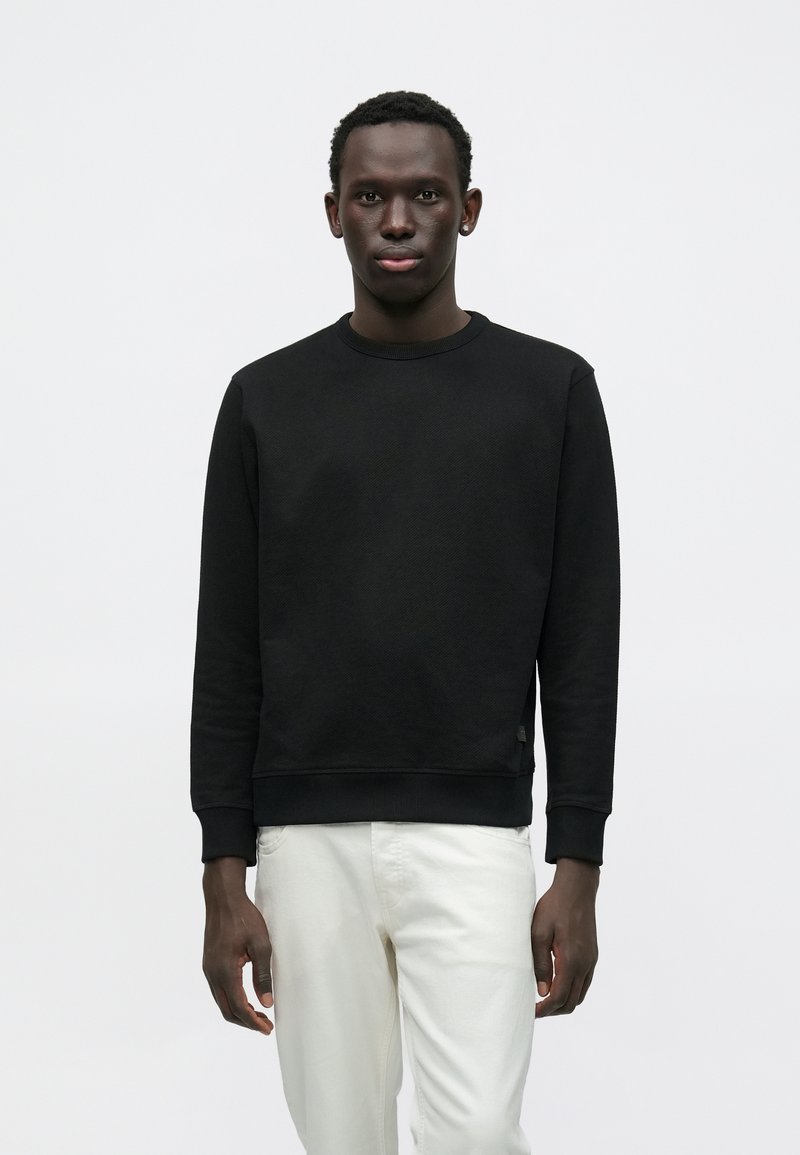 Μαύρο φούτερ με ριμπ λαιμόκοψη και μανσέτες, με υφή υφάσματος και άνετη εφαρμογή. Συνδυάζεται με λευκό παντελόνι.