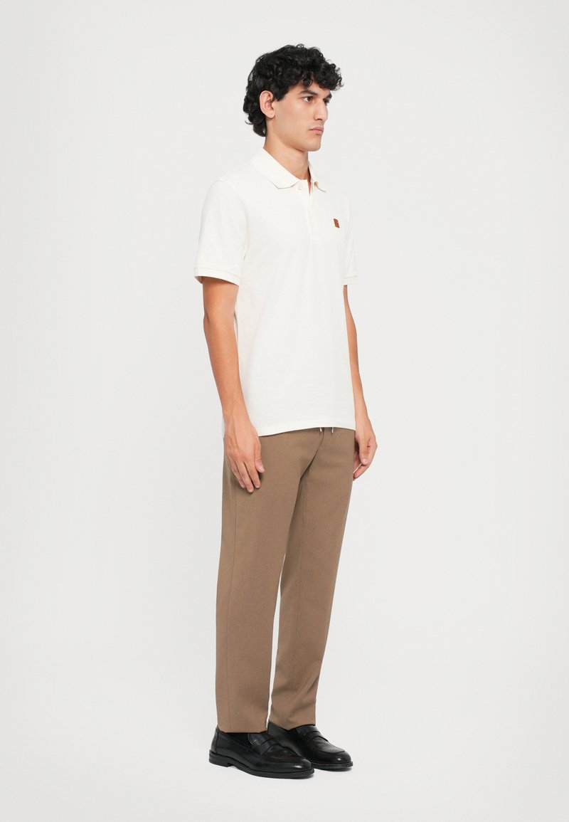 Hellbeige Poloshirt mit kurzen Ärmeln, Kragen und einem kleinen gestickten Logo. Kombiniert mit schmal geschnittenen braunen Hosen und schwarzen Schuhen.
