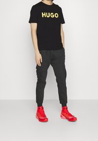 Camiseta de algodón negra con el logo amarillo "HUGO", combinada con pantalones cargo gris oscuro y zapatillas deportivas rojas brillantes con superficies texturizadas.