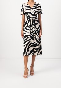 Midi šaty se zebrovým potiskem v béžové a černé, s překládaným designem, krátkými rukávy, páskem v oblasti pasu a rozšířeným lemem. Nosené s nazouvacími béžovými podpatky.