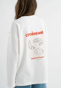 Witte oversized sweatshirt met een grafisch ontwerp van drie croissants in lijnstijl. De tekst luidt "croissant" en "made in France" in het rood.