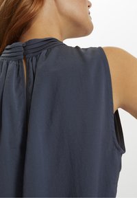 Námořnická modrá bezramenná blůza s dírkou na zádech, jemnými sklady a hladkou texturou látky. Knoflíky jsou viditelné na výstřihu.