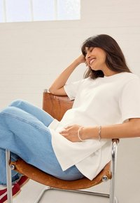 Wit T-shirt met korte mouwen en een relaxte pasvorm, gecombineerd met lichtblauwe denim jeans. Het model zit op een bruine leren stoel en draagt een zilveren armband.
