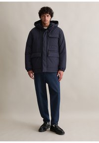 Granatowa ocieplana kurtka z kapturem, cztery przednie kieszenie, zamek błyskawiczny, elastyczne mankiety, w połączeniu z ciemnoniebieskimi spodniami dżinsowymi i czarnymi butami.